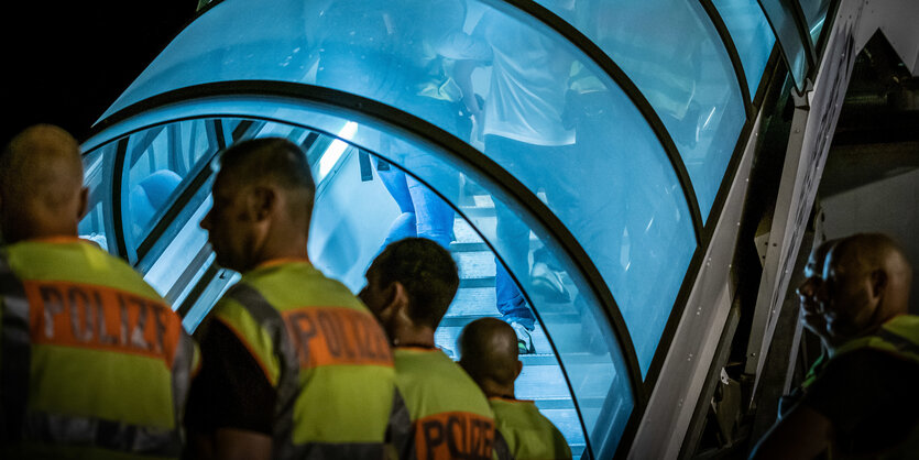 Ein Mann wird bei Dunkelheit eine blau beleuchtete Flugzeugtreppe hinaufgeführt, im Vordergrund steht Sicherheitspersonal