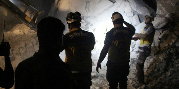 Drei Männer stehen mit dem Rücken zur Kamera in einem dunkelen Raum voller Trümmer. Einer von ihnen hält eine Taschenlampe gegen die Rückwand.