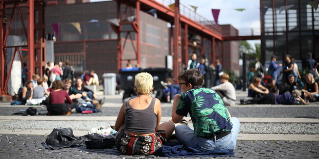 Zwei Menschen sitzen mit dem Rücken zur Kamera auf dem Kopfsteinpflaster, die Sonne scheint. Im Hintergrund sitzen weitere Menschen auch auf dem Boden