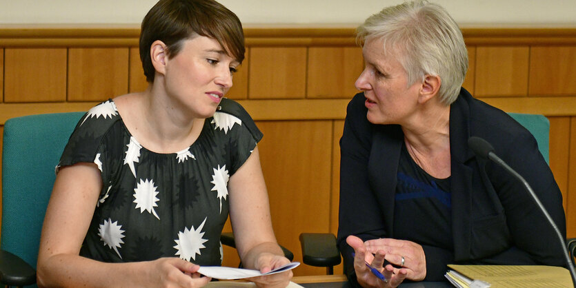 Zwei Frauen sitzen an einem Tisch in einem Gerichtssaal. Sie sind einander zugewandt und unterhalten sich.