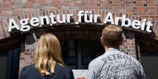 Eine Frau mit langen blonden Haaren und ein Mann mit kurzen braunen Haaren stehen vor dem Eingang zu einer Agentur für Arbeit