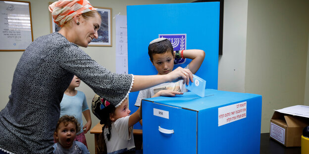 Eine Frau wirft einen Stimmzettel in eine Wahlurne. Kinder stehen um sie herum.
