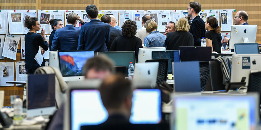 In einem großen Raum stehen einige Personen vor einer Wand mit Zeitungsseiten, andere sitzen an ihren Arbeitsplätzen und gucken auf einen Bildschirm