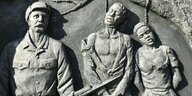 Ein Denkmal in Windhoek, Namibia, zeigt einen weißen Kolonialisten mit Uniform und Gewehr. Neben ihm stehen zwei schwarze ausgemergelte Personen mit einem Strick um den Hals.