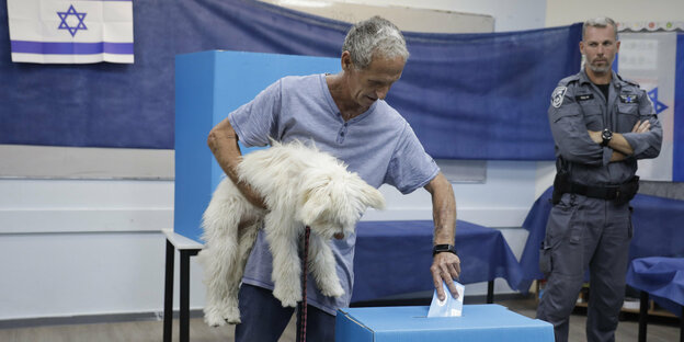 Ein Mann wirft seinen Wahlzettel in die Urne, in der rechten Hand trägt er einen zotteligen weißen Hund. Im Hintergrund steht ein israelischer Polizist
