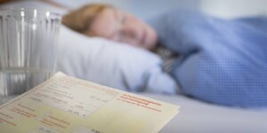 Eine Frau liegt krank im Bett, im Vordergrund ist ein Krankmeldungsschein zu sehen