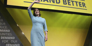 Die Anführerin der Liberaldemokraten Lib-Dem, Jo Swinson, winkt bei einer Parteiveranstaltung. Sie trägt ein langes Kleid und steht auf einer Bühne. Hinter ihr ein großes gelbes Plakat der Lib-Dems. Darauf steht: "Liberal Democrats. Demand Better"