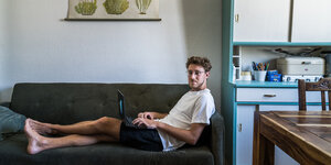Ein junger Mann liegt mit Laptop auf dem Schoß auf einer Couch