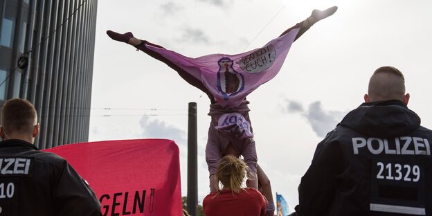 Eine Frau in rosa Hosen macht einen Handstand auf den Händen einer anderen. An ihrem Bein hängt ein Schriftzug auf dem "Verpisst euch" steht.