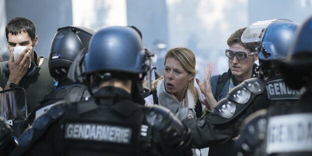 Menschen stehen vor Polizisten, einer hält sich ein Taschentuch vor den Mund, ein anderer trägt eine Schutzbrille