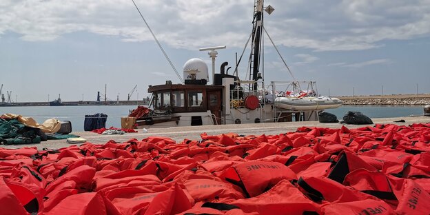 Rote Rettungswesten sind auf einem Pier aufgestapelt, dahinter liegt ein Rettungsschiff