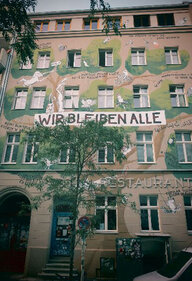 Das sechsstöckige Haus an der Waldermarstraße mit einer bemalten Fassade. Die Malerei zeigt einen großen Baum, der sich um die Fenster schlängelt. Dazwischen sind Tiere gemalt. In der Mitte steht: "Wir bleiben alle."