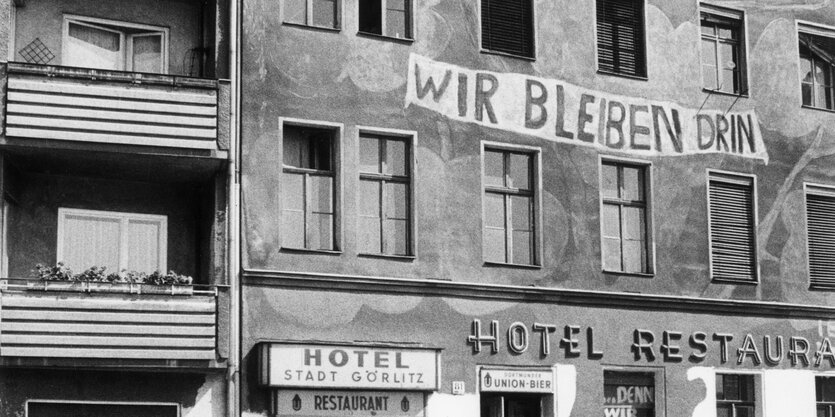 Ein Archivbild des Hauses in schwarz weiss. An der Fassade steht: "Wir bleiben drin."