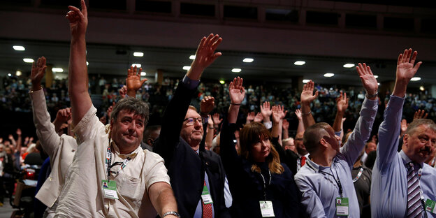 Menschen in einer vollen Kongresshalle heben jeweils eine Hand Menschen in einer vollen Kongresshalle heben jeweils eine Hand