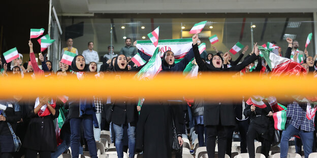 Iranische weibliche Fußballfans jubeln mit Flaggen, hinter einem gelben Balken