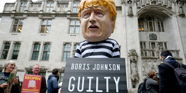 Ein Mann, der eine riesige Boris Johnson-Maske und Sträflingskleidung trägt, hält ein Schild, auf dem steht «Boris Johnson GUILTY»