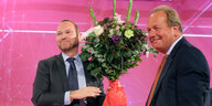 Frank Werneke hält einen Blumenstrauß hoch, neben ihm Frank Bsirske