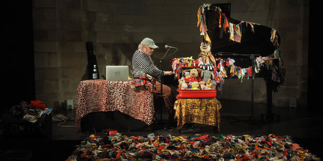 Musiker Charlemagne Palestine sitzt auf der Bühne inmitten von bunten Kuscheltieren und einem Laptop an einem Flügel und singt