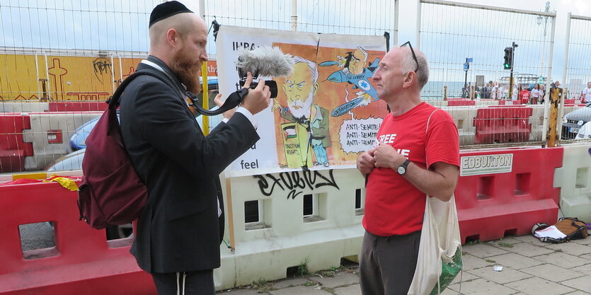 Zwei Männer vor einem Plakat. Einer von ihnen trägt eine Kippa