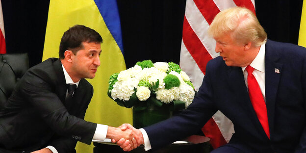 Ukraines Präsident Wolodimir Selenski und Donald Trump schütteln sich die Hände, im Hintergrund Fahnen und ein Blumenstrauß
