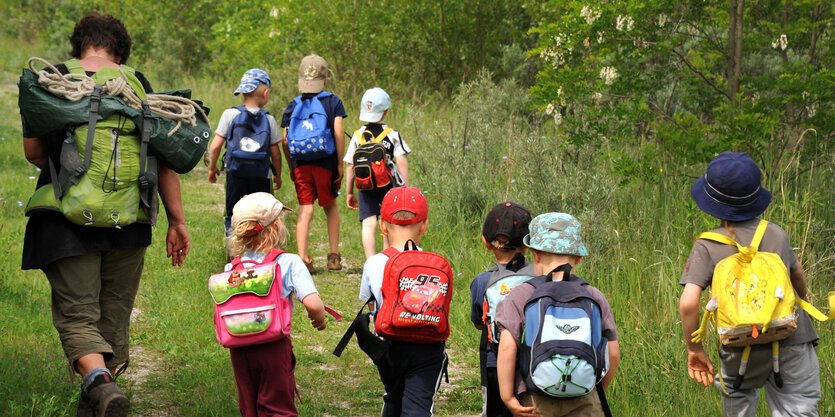 Eine Erzieherin läuft mit vielen Kita-Kindern über eine Wiese. Sie tragen Rucksäcke