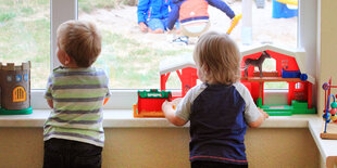 Zwei kleine Kinder schauen neben buntem Spielzeug aus dem Fenster