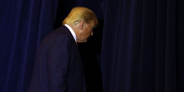 Trump verlässt das Podium einer Pressekonferenz. In seinem dunkelblauen Anzug geht er vor dem gleichfarbigen Vorhang beinahe unter.