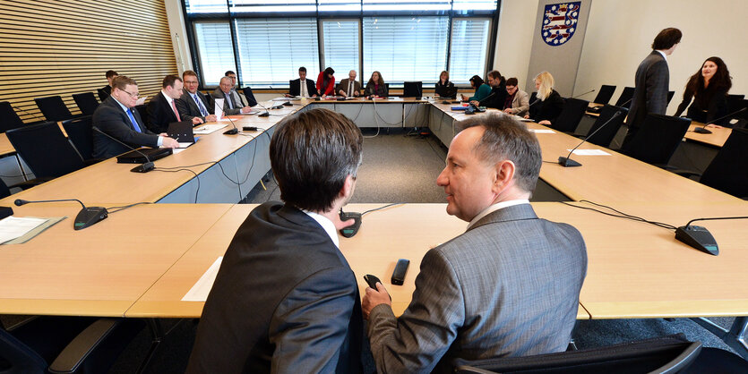 zwei diskutierende Menschen in einem Sitzungssaal im Vordergrund, weitere Menschen im Hintergrund.