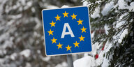 Ein blaues Schild mit einem weißen A in der Mitte und den EU-Sternen drumherum steht vor einem verschneiten Hintergrund.