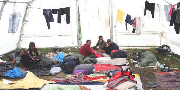 Menschen sitzen in einem großen Zelt, um sie herum liegen und hängen Kleidungsstücke