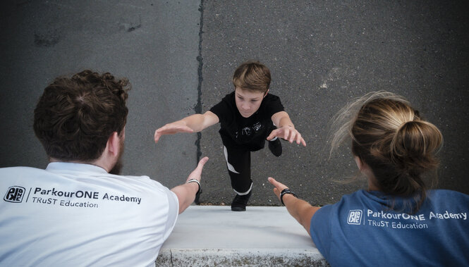 Zwei Erwachsene helfen einem Kind, eine Mauer hoch zu kommen - ein Foto aus der Parkour-Schule am Berliner Veledrom