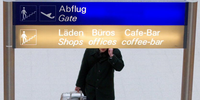 GHinweiosschild an einbem FLughafen. Dahinter ein Fluggast mit Rollkoffer und Mobiltelefon