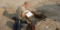 Ein Mann sitzt in einer Sandgrube und zeichnet einen Tonkrug ab