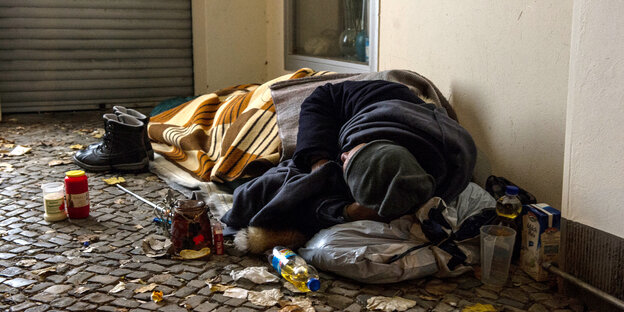 Ein Wohnungsloser liegt auf einer Matte auf dem Boden, in Decken eingewickelt.
