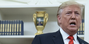 Donald Trump mit missmutigem Gesichtsausdruck vor einem Regal, in dem ein goldener Pokal und Bücher stehen