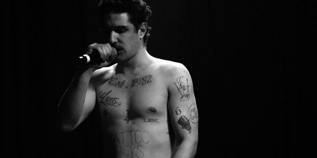 Der Sänger Joey Bargeld steht mit nacktem Oberkörper angeleuchtet im Dunkeln und hält ein Mikrophon in der Hand