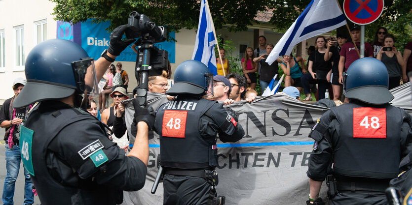 Teilnehmer einer Gegendemonstration mit Transparenten stehen hinter Einsatzkräften der Polizei, die mit Kameras filmt