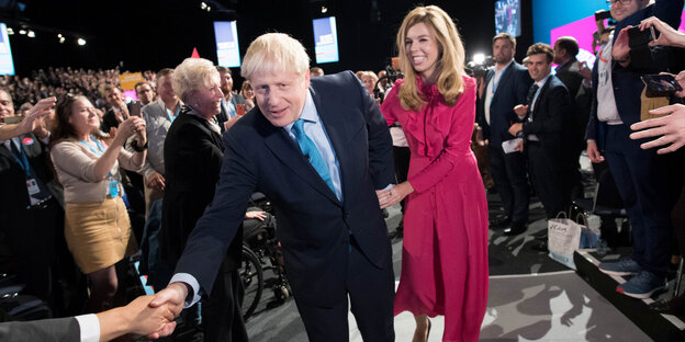 Boris Johnson im Anzug, händeschüttelnd in einer Menschenmenge, neben ihm eine Frau in einem pinken Kleid