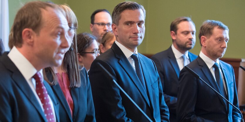 Drei Männer im Anzug, dahinter weitere Menschen