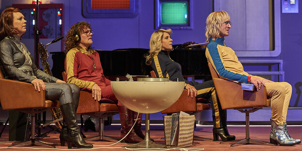 Vier Schauspieler sitzen in trashigen Kostümen auf Stühlen auf einer Bühne