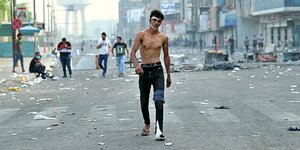 ein Mann mit nacktem Oberkörper und zerrissenen Jeans läuft eine Straße entlang, auf der Ausschreitungen stattgefunden haben. Im Hintergrund vermummte Demonstranten