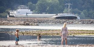 an einem Flussufer baden Menschen, im Hintergrund fährt ein Frachtschiff