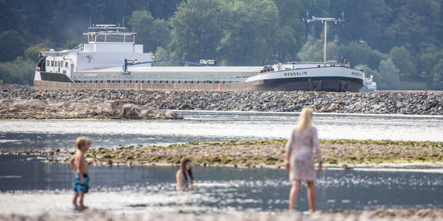 an einem Flussufer baden Menschen, im Hintergrund fährt ein Frachtschiff
