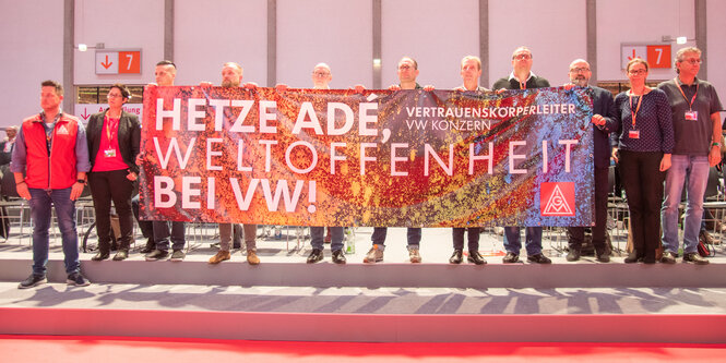 Männer und Frauen stehen auf einer Bühne und halten ein Banner mit der Aufschrift "Hetze adé, Weltoffenheit bei VW"