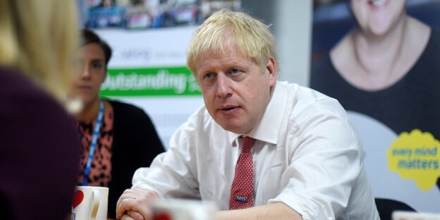 Boris Johnson sitzt ohne Jackett an einem Tisch und faltet die Hände