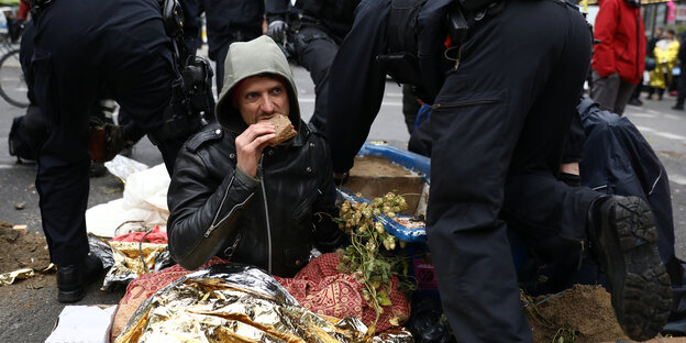 Ein Mensch sitzt an einem Gegenstand festgekettet auf der Straße und isst ein Pausenbrot