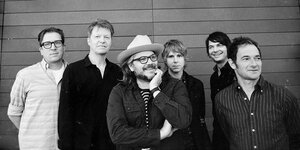 Ein schwarzweißes Foto, sechs Männer, milde bis freundlich lächelnd, posieren vor einer Wand. Ein trägt Hut (Wilco).