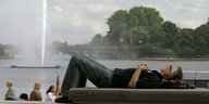Ein Mann liegt auf einer Bank, hinter ihm ein See mit Fontäne.