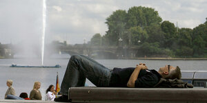 Ein Mann liegt auf einer Bank, hinter ihm ein See mit Fontäne.