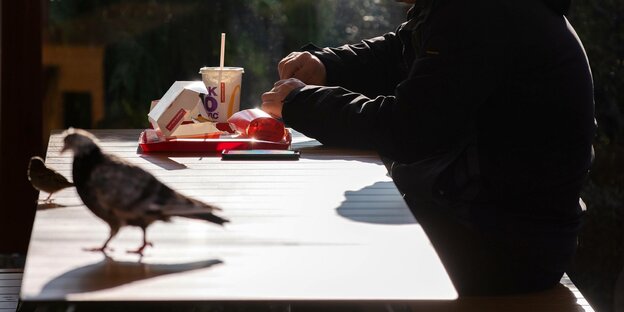 Taube auf einem Tisch an dem ein MAnn vor einem Tablet mit Fastfood sitzt